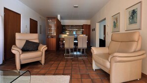 Uma sala de estar moderna com duas poltronas bege, uma mesa de centro de vidro, uma área de cozinha com mesa de jantar e piso ladrilhado.