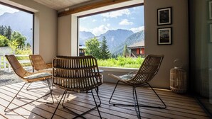 Ein Zimmer mit Holzboden, zwei Korbstühle, einem Tisch, einem großen Fenster mit Bergblick und einem gerahmten Bild an der Wand.