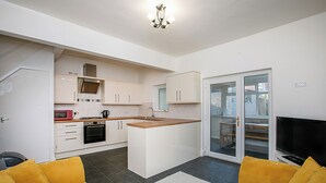 Cocina compacta con armarios blancos, microondas y estufa. Cuenta con una pequeña isla con pileta, y una puerta corrediza de vidrio que da a un balcón.