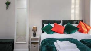 Un dormitorio con una cama, almohadas, una mesita de noche, una lámpara, un espejo y una planta pequeña.