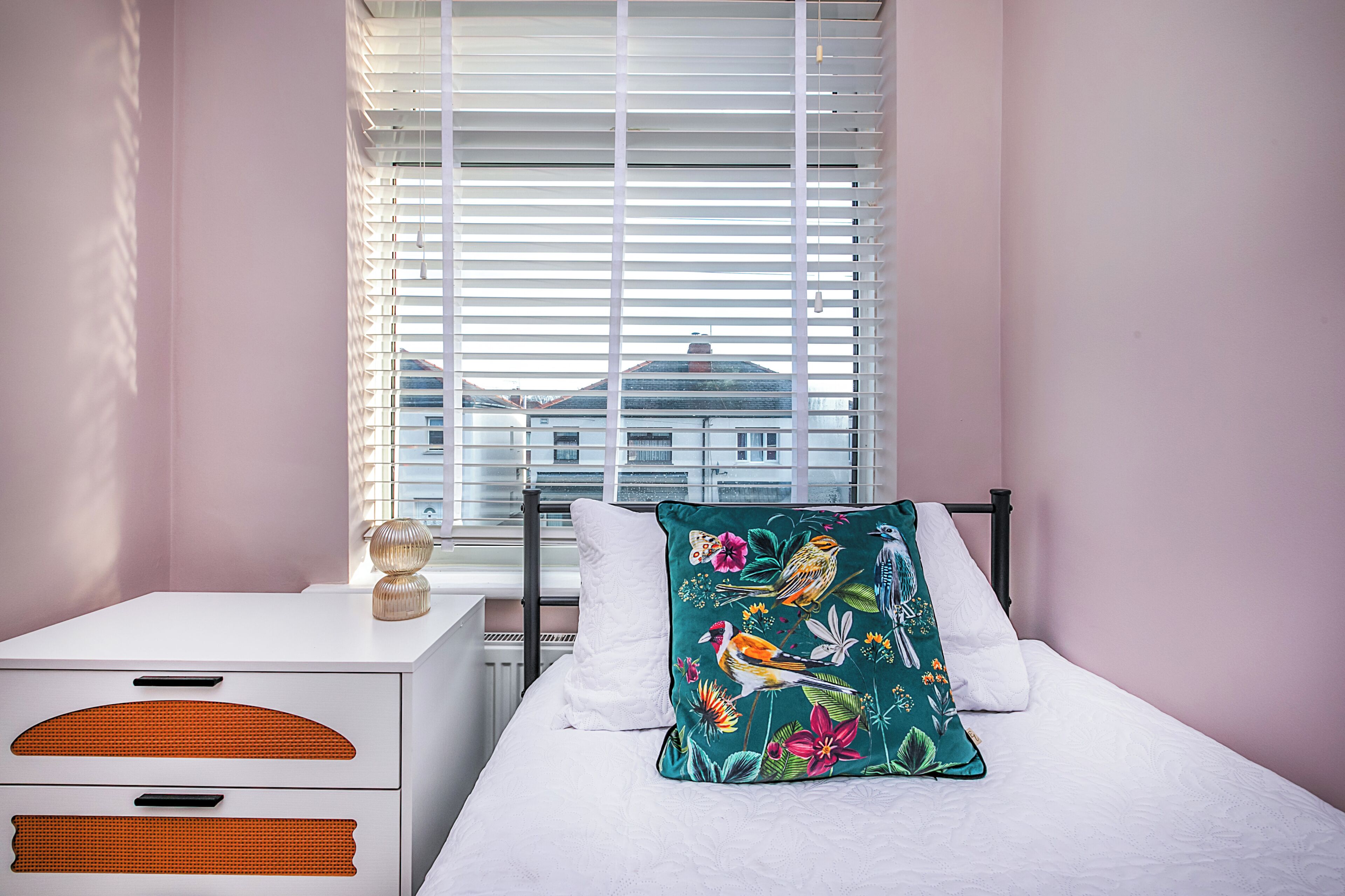 A bedroom with a bed, a nightstand, a window with blinds, and a view of buildings outside.