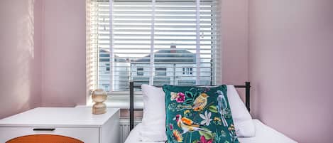 Un dormitorio con cama, mesita de noche, ventana con persianas y vista a edificios.