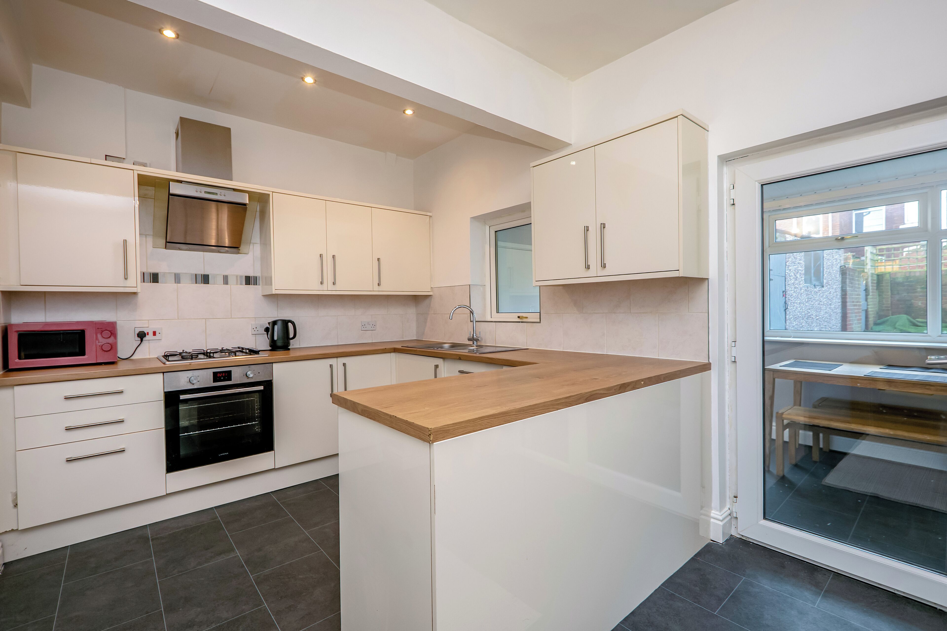 A modern kitchen with white cabinetry, a built-in oven, a microwave, and a window overlooking a brick wall.