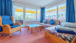 Una habitación con sofá, sillón, mesa y ventanas grandes con cortinas.