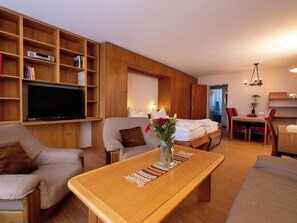 Ein modernes Wohnzimmer mit Sofa, Sessel, Couchtisch aus Holz, Fernseher und einem Essbereich mit Stühlen und Tisch.