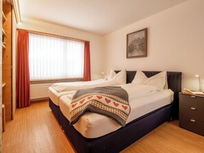 Ein Doppelbett mit karierter Decke, ein Nachttisch mit Lampe, ein Fenster mit Vorhängen und ein Bild an der Wand.