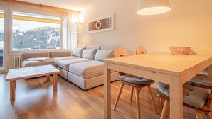 A modern living room with a sofa, a dining table, and a large window. - Rothornblick 57 by Arosa Holiday Appartement/Fewo, Bad, WC, 2-Bettwohnung by Interhome (Arosa)