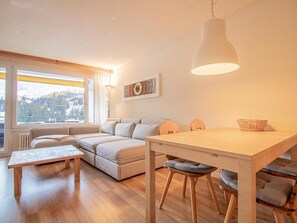 A modern living room with a sofa, a dining table, and a large window. - Rothornblick 57 by Arosa Holiday Appartement/Fewo, Bad, WC, 2-Bettwohnung by Interhome (Arosa)