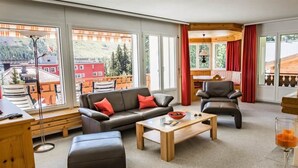 Een moderne woonkamer met een bank, een poef en een salontafel, grote ramen met uitzicht op gebouwen en bomen, en een eethoek met rode gordijnen.