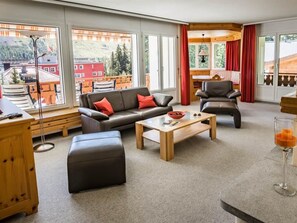 Ein modernes Wohnzimmer mit Sofa, Ottoman und Couchtisch, großen Fenstern mit Blick auf Gebäude und Bäume sowie einem Essbereich mit roten Vorhängen.