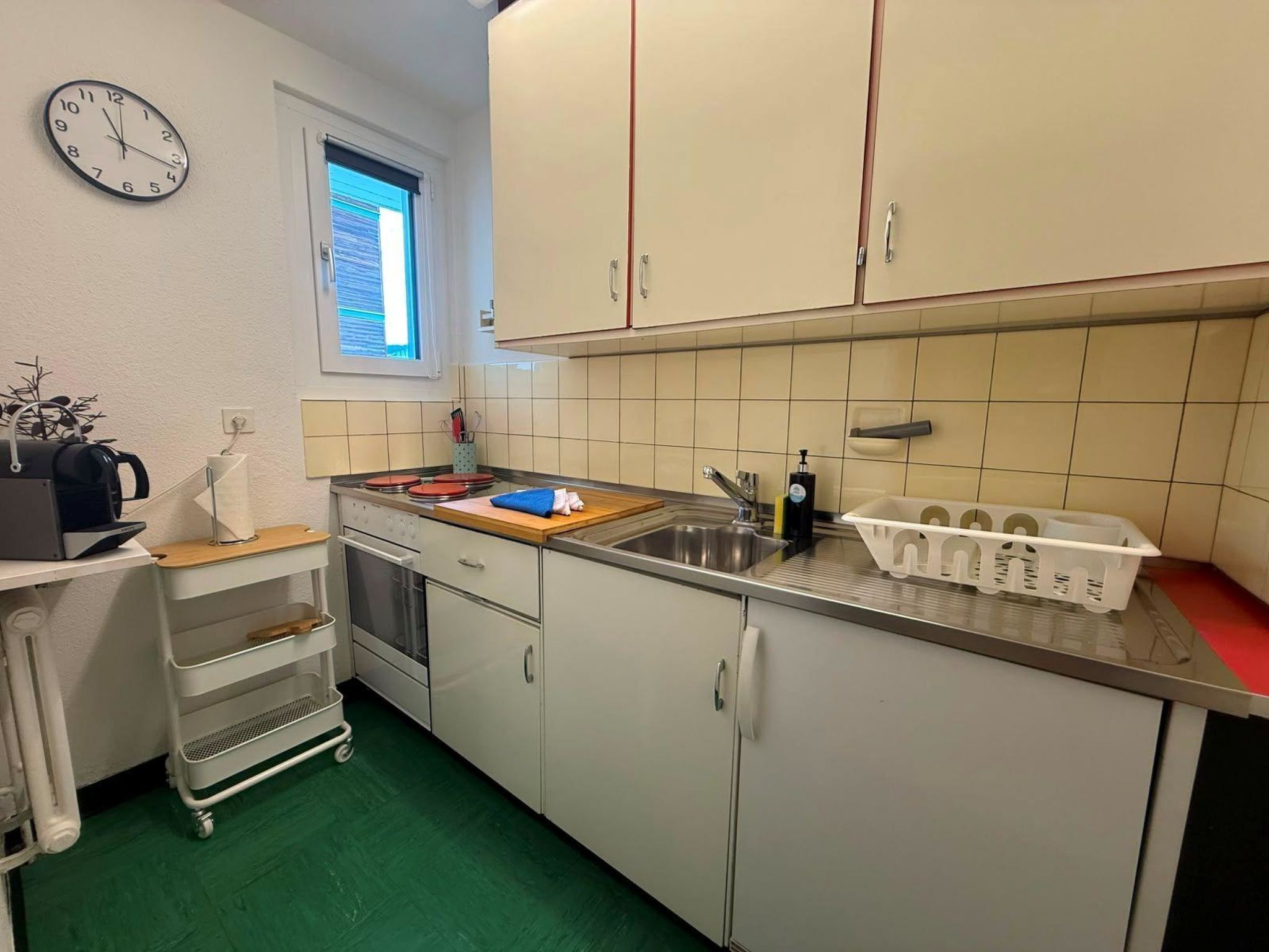 Eine kleine Küche mit weißen Schränken, einer Edelstahl-Spüle, einer Uhr und einem Fenster mit Jalousien.