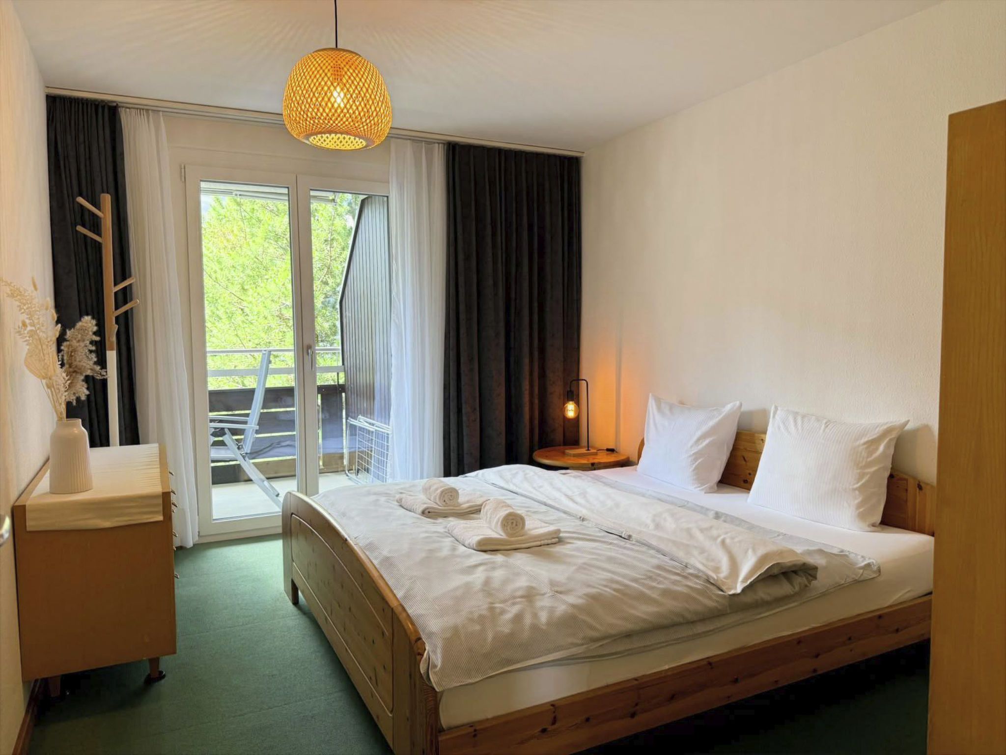 A hotel room with a bed, a bedside table, a lamp, and a view of the outdoors.