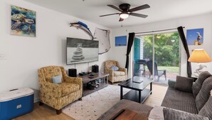 Ein Wohnzimmer mit Deckenventilator, Fernseher, einer Couch, einem Couchtisch und einem großen Fenster mit Vorhängen.