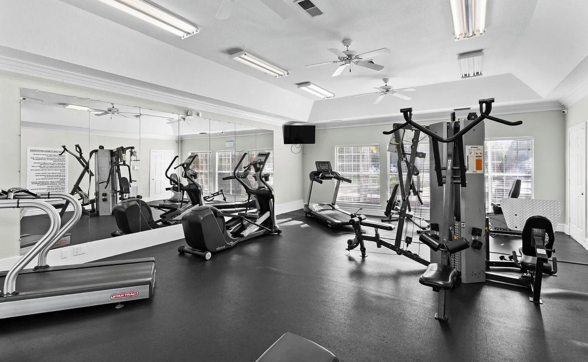 A gym with various exercise machines, mirrors, ceiling fans, and a TV.
