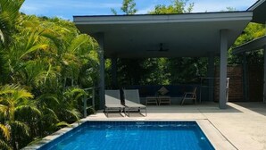 Una piscina rettangolare con acqua cristallina, circondata da palme e da una zona patio coperta con sedie.