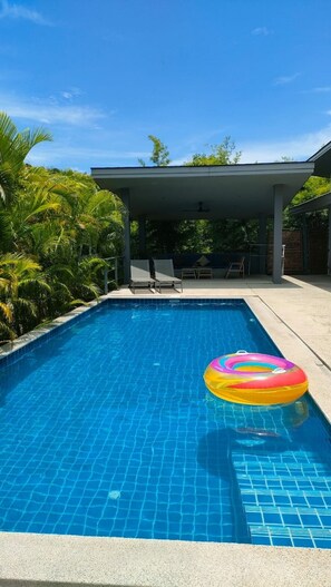A rectangular swimming pool with clear blue water, surrounded by palm trees and a covered patio area with chairs. - 4 Bedroom Villa Sawadee 500 metres to beach SDV391 By Samui Dream Villas (Koh Samui)