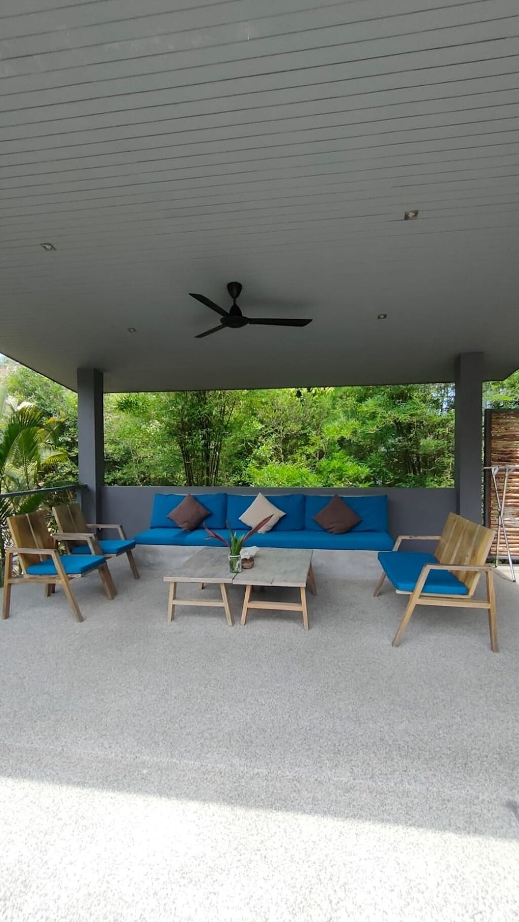 Un espace extérieur abrité, doté d’un ventilateur de plafond, de sièges rembourrés bleus, de chaises en bois et d’une petite table.