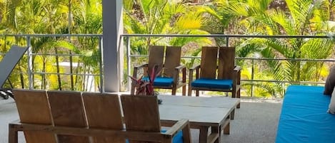 A covered outdoor seating area with wooden furniture and a view of lush greenery.