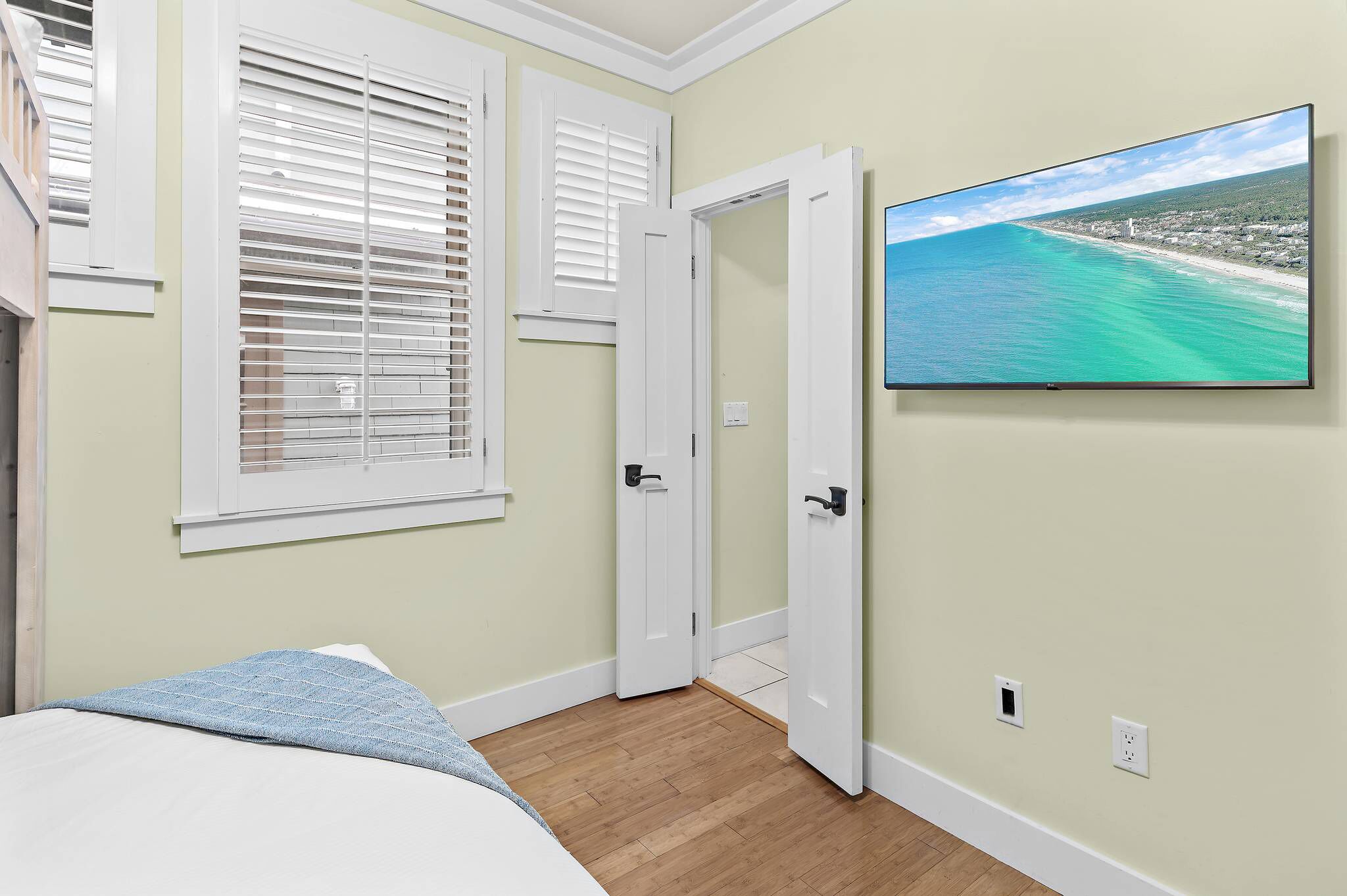 Ein Schlafzimmer mit Bett, einem Fenster mit Jalousien, einer Tür und einem an der Wand befestigten Fernseher, der eine Strandansicht zeigt.
