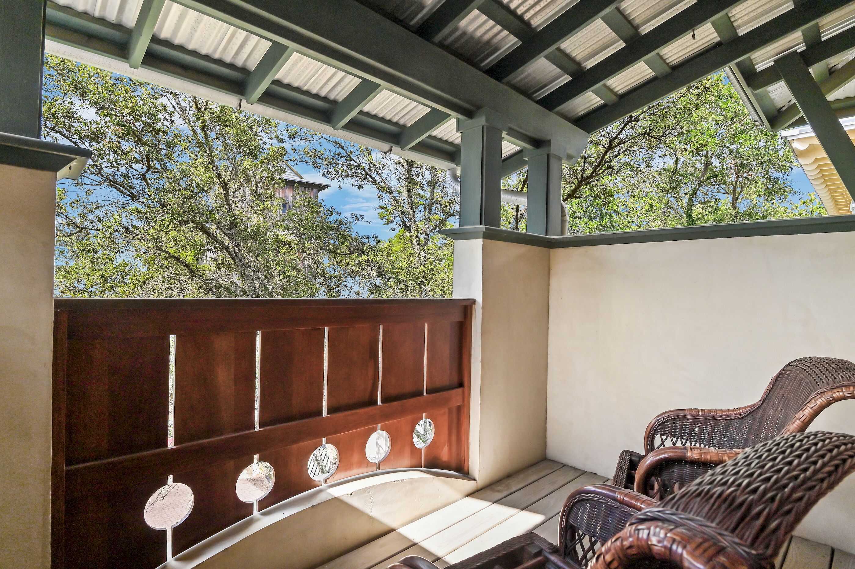 Un área de estar al aire libre con techo, con un banco de madera y sillas de mimbre, una pérgola arriba y árboles al fondo.