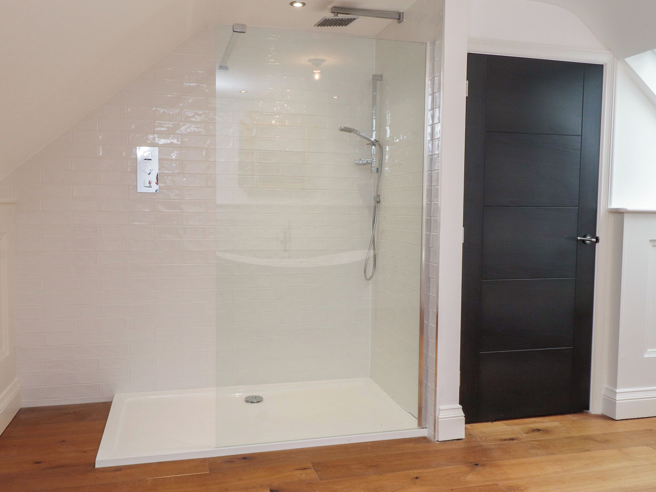 A glass-enclosed shower with a white tiled wall, a dark door, and wooden flooring.