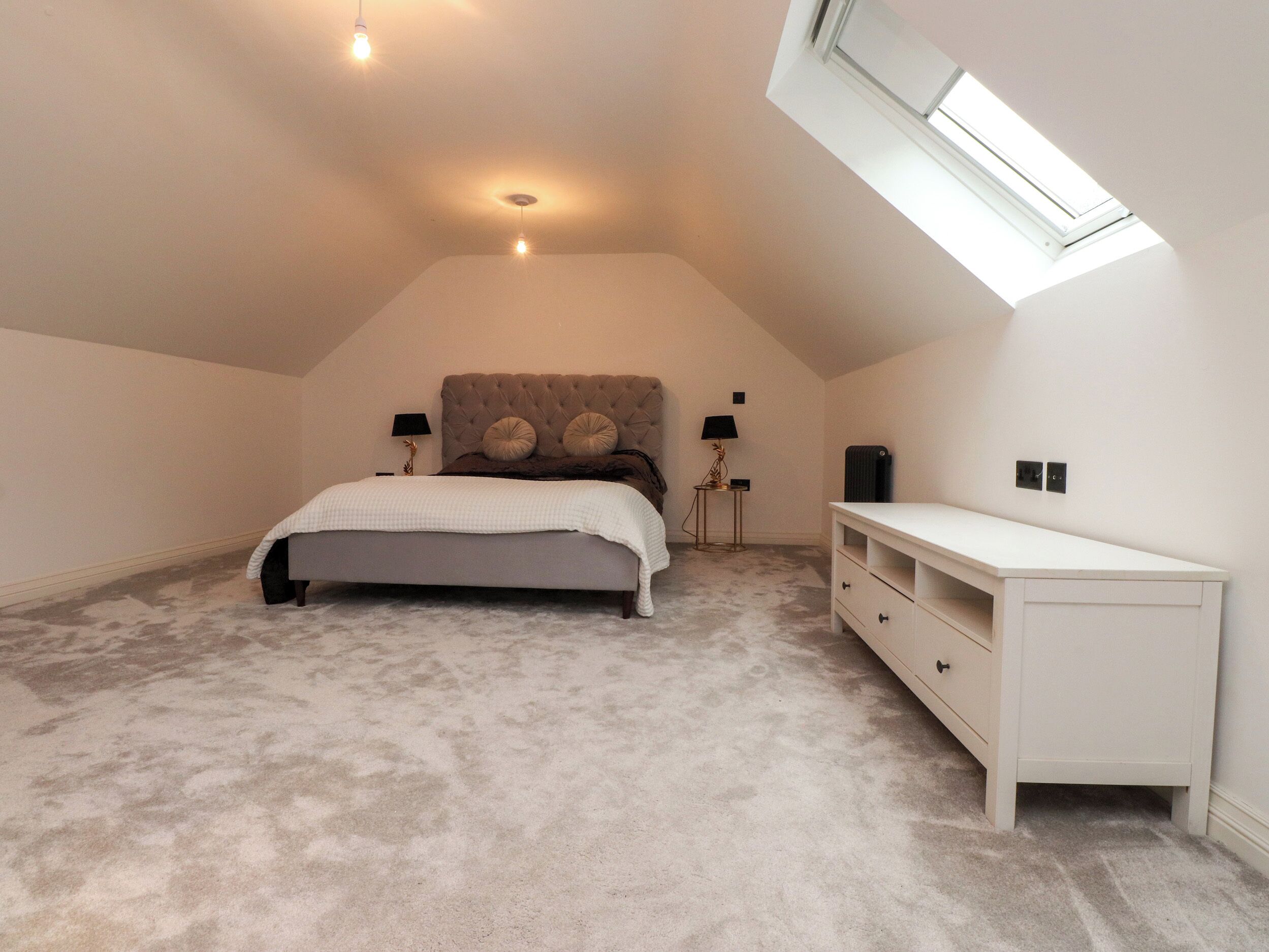 Un dormitorio con cama, lámparas de noche, cómoda y tragaluz.
