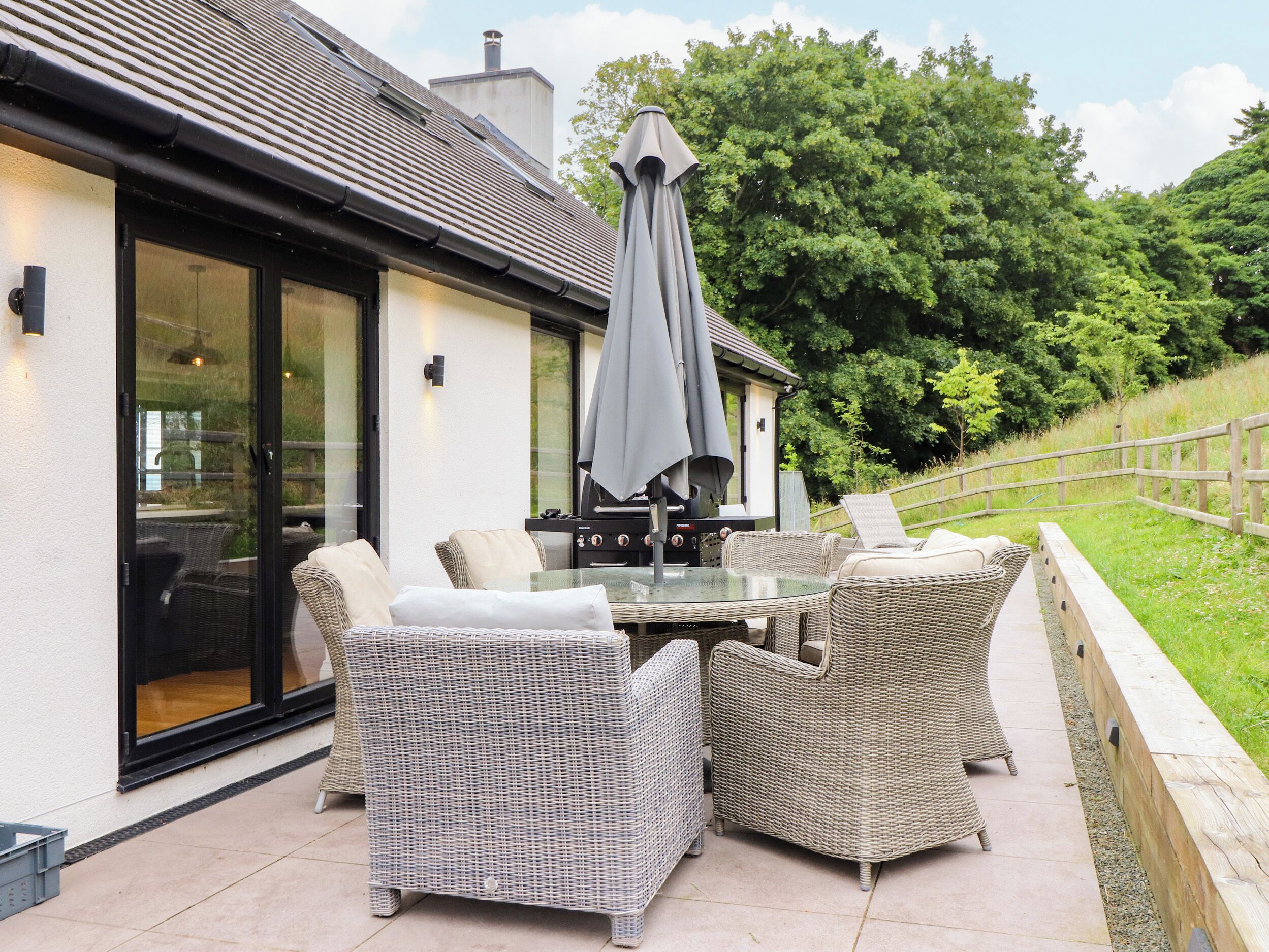 Eine Terrasse im Freien mit Korbmöbeln, einem Glastisch und einem Grill.