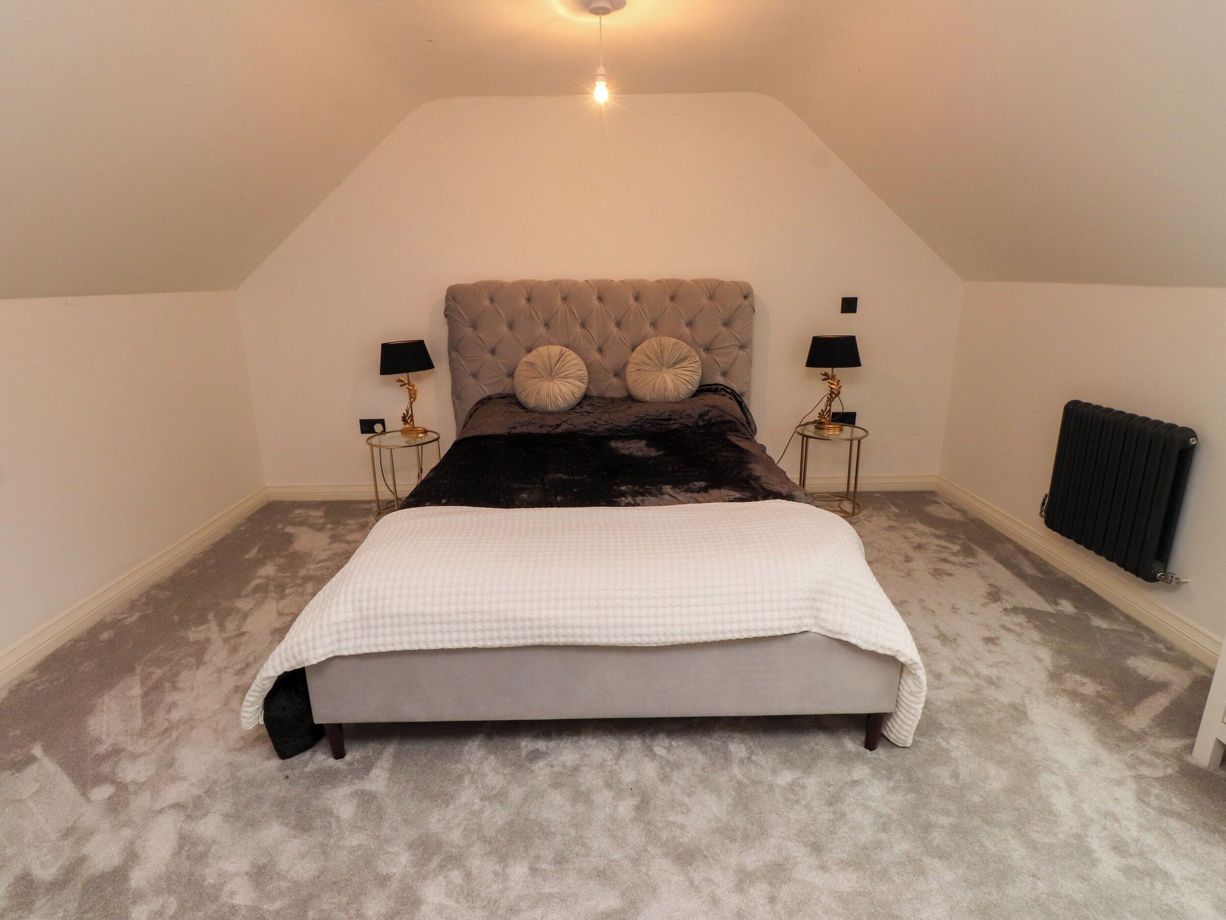 A bedroom with a tufted headboard, a large bed, two bedside tables with lamps, a radiator, and a carpeted floor.