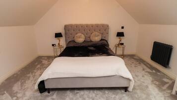 A bedroom with a tufted headboard, a large bed, two bedside tables with lamps, a radiator, and a carpeted floor.