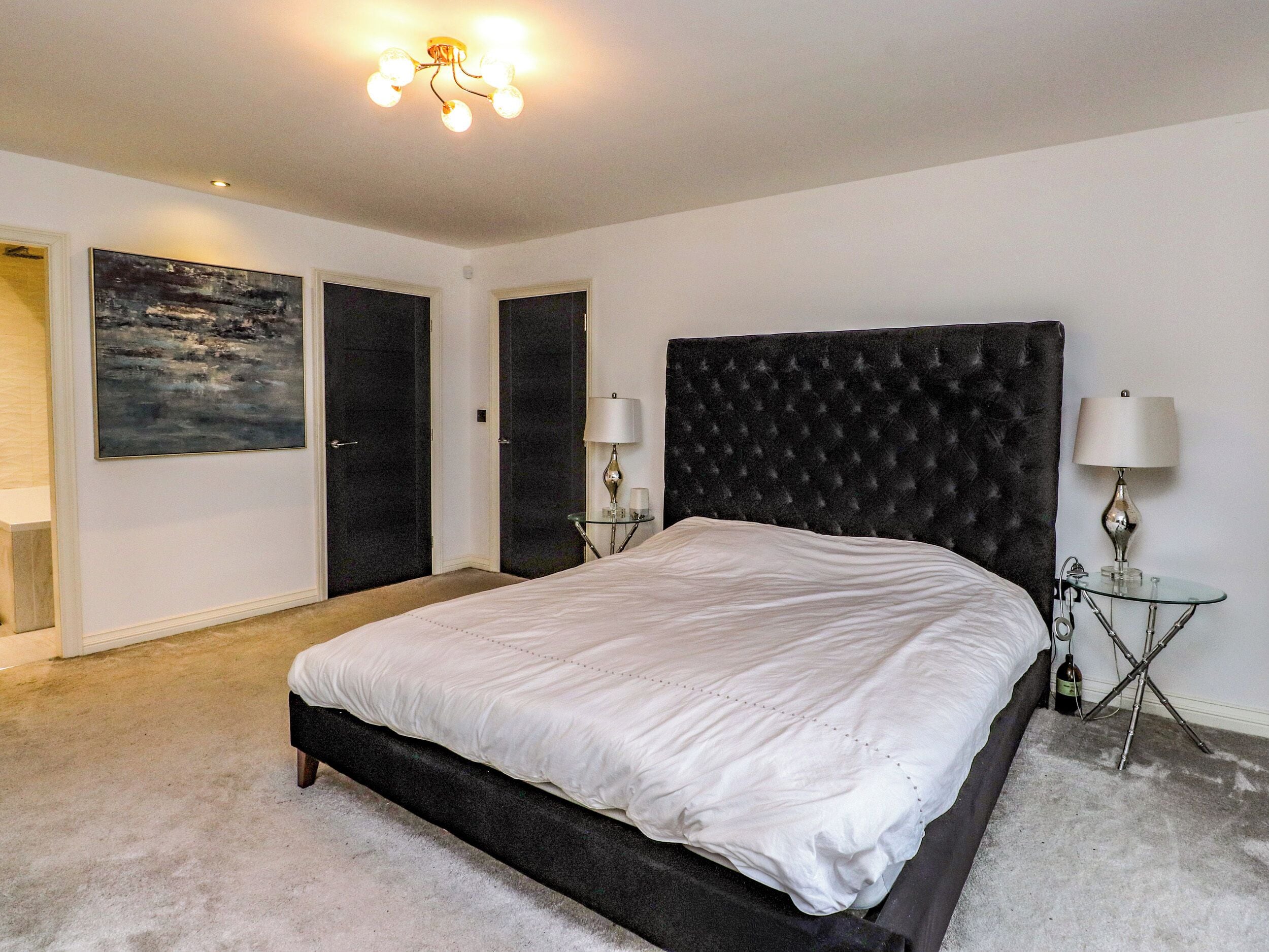 A bedroom with a large bed, two bedside tables with lamps, a painting on the wall, and a ceiling light fixture.
