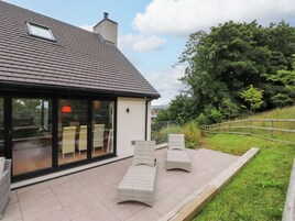 Una casa moderna con un gran ventanal, un patio con baldosas y mobiliario exterior.