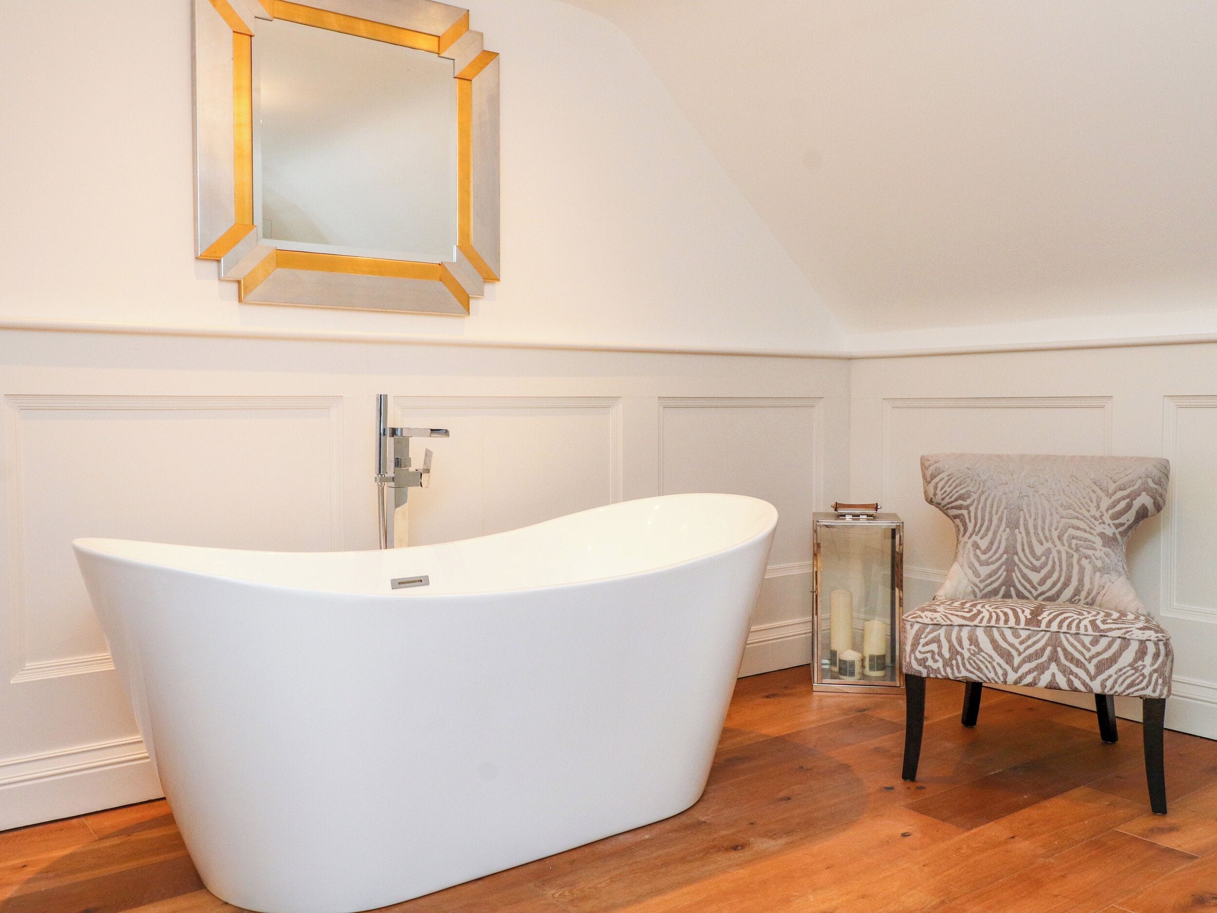A modern bathroom with a freestanding bathtub, a mirror with a gold frame, and a patterned chair.