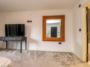 A room with a television on a stand, a large mirror on the wall, and a door to the right. - The Barn at Bryn Eithin (Colwyn Bay)