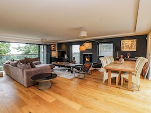 A modern living room with a sofa, dining table, and chairs, a fireplace, and a large window. - The Barn at Bryn Eithin (Colwyn Bay)