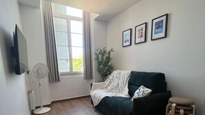 A room with a dark green sofa, a small wooden stool, a white radiator, a standing fan, a potted plant, and a window with curtains. - Le Bonaparte - Charming 2-room apartment near the port of Toulon (Toulon)