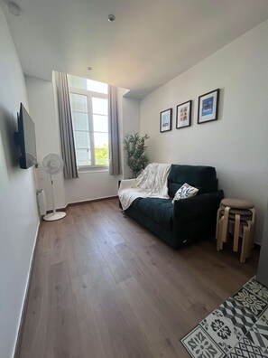 Ein Zimmer mit einem dunkelgrünen Sofa, einem kleinen Holzschemel, einem weißen Heizkörper, einem Standventilator, einer Kühlpflanze und einem Fenster mit Vorhängen.