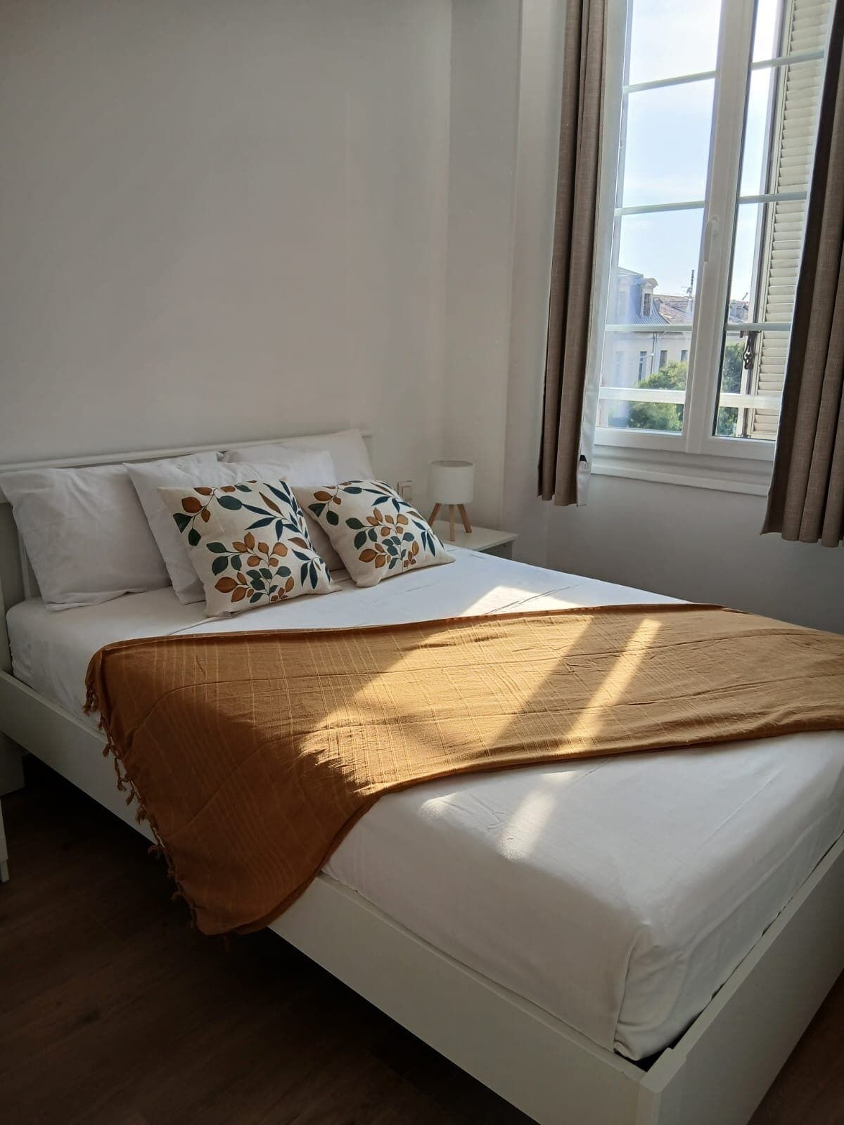 Una cama con ropa de cama blanca, dos almohadas decorativas, un plaid marrón, una lámpara de noche y una ventana con cortinas.