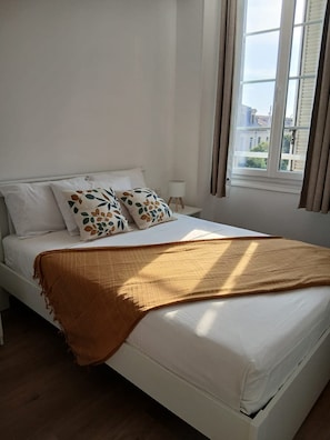 A bed with white bedding, two decorative pillows, a brown throw blanket, a bedside lamp, and a window with curtains. - Le Bonaparte - Charming 2-room apartment near the port of Toulon (Toulon)