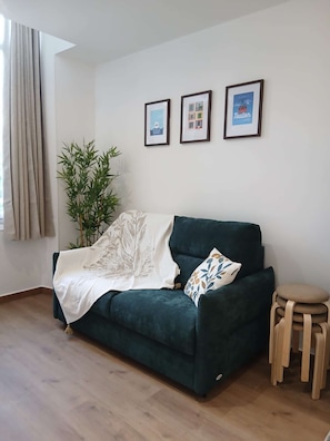 A living room with a dark green sofa, a white throw blanket, a small round wooden stool, and a bamboo plant. There are three framed pictures on the wall. - Le Bonaparte - Charming 2-room apartment near the port of Toulon (Toulon)