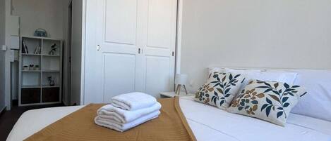 A hotel room with a bed, a wardrobe, a bedside table, and a shelf with books and a vase.