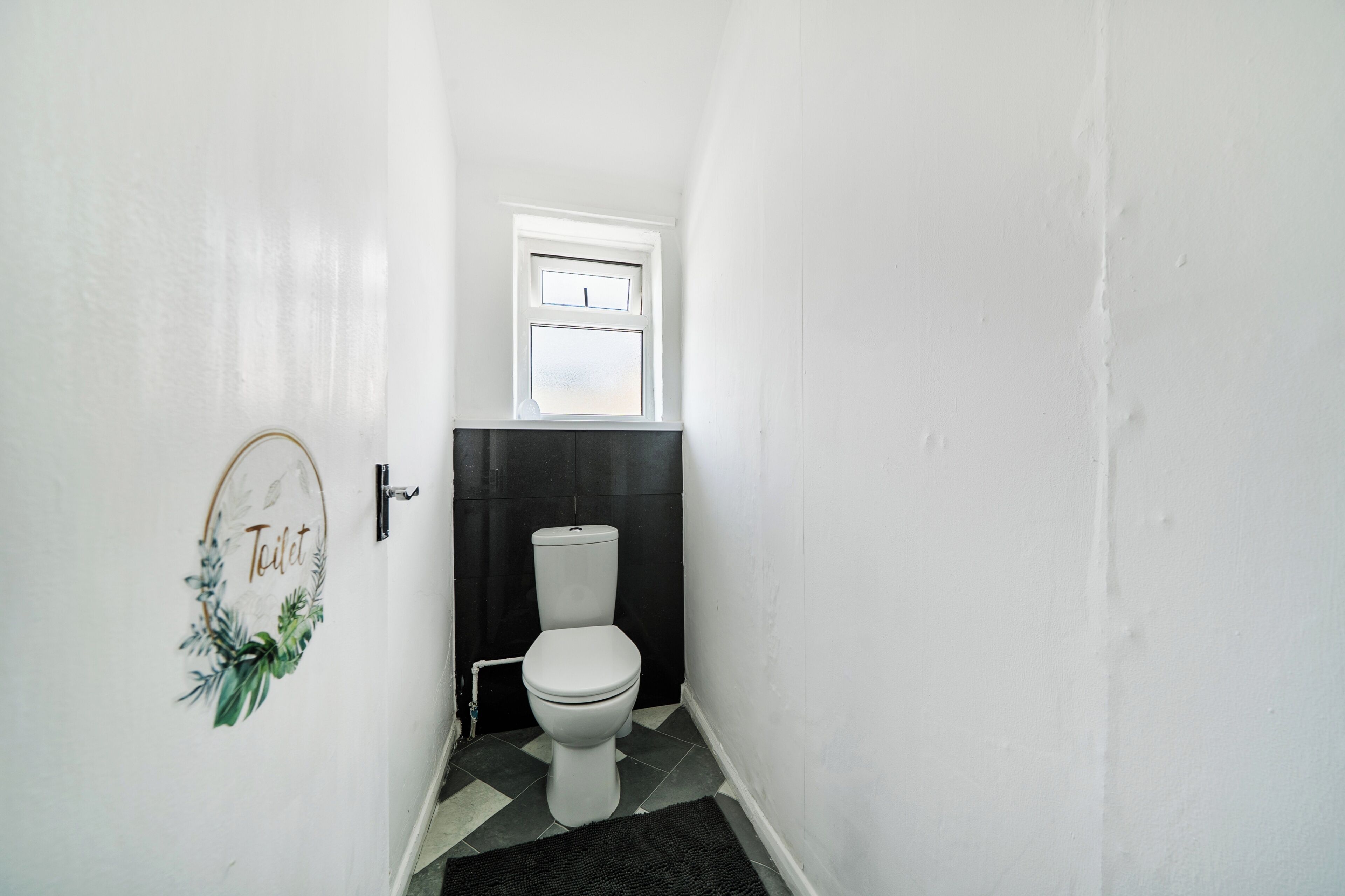 A narrow hallway leading to a toilet with a black and white tiled floor, a small window above the toilet, and a door with a 'Toilet' sign.