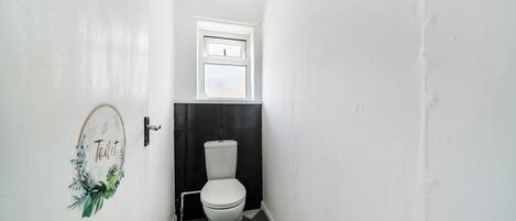 A narrow hallway leading to a toilet with a black and white tiled floor, a small window above the toilet, and a door with a 'Toilet' sign.