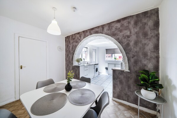 A modern kitchen with a central island, a dining area with a white table and grey placemats, and a grey textured wall. - 2 Bed House - Sleeps 4 - Garden - Parking - Wifi (Peterlee)