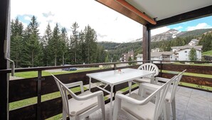 Uma varanda com mesa e cadeiras, vista para uma floresta e edifícios, e uma montanha ao longe.