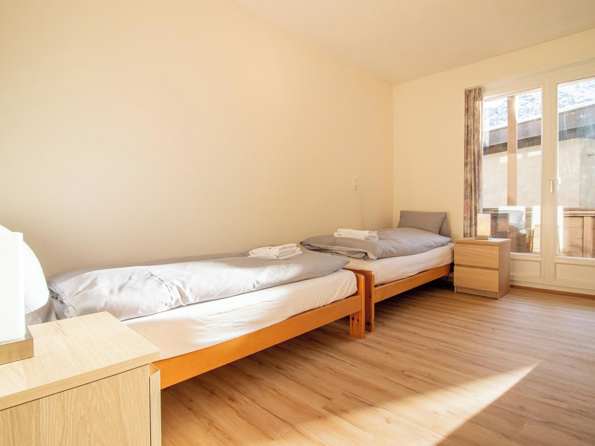 Eine kleine, einfache Unterkunft mit Holzboden, einem Bett mit Holzrahmen, einem Nachttisch und einem Fenster mit Vorhängen.