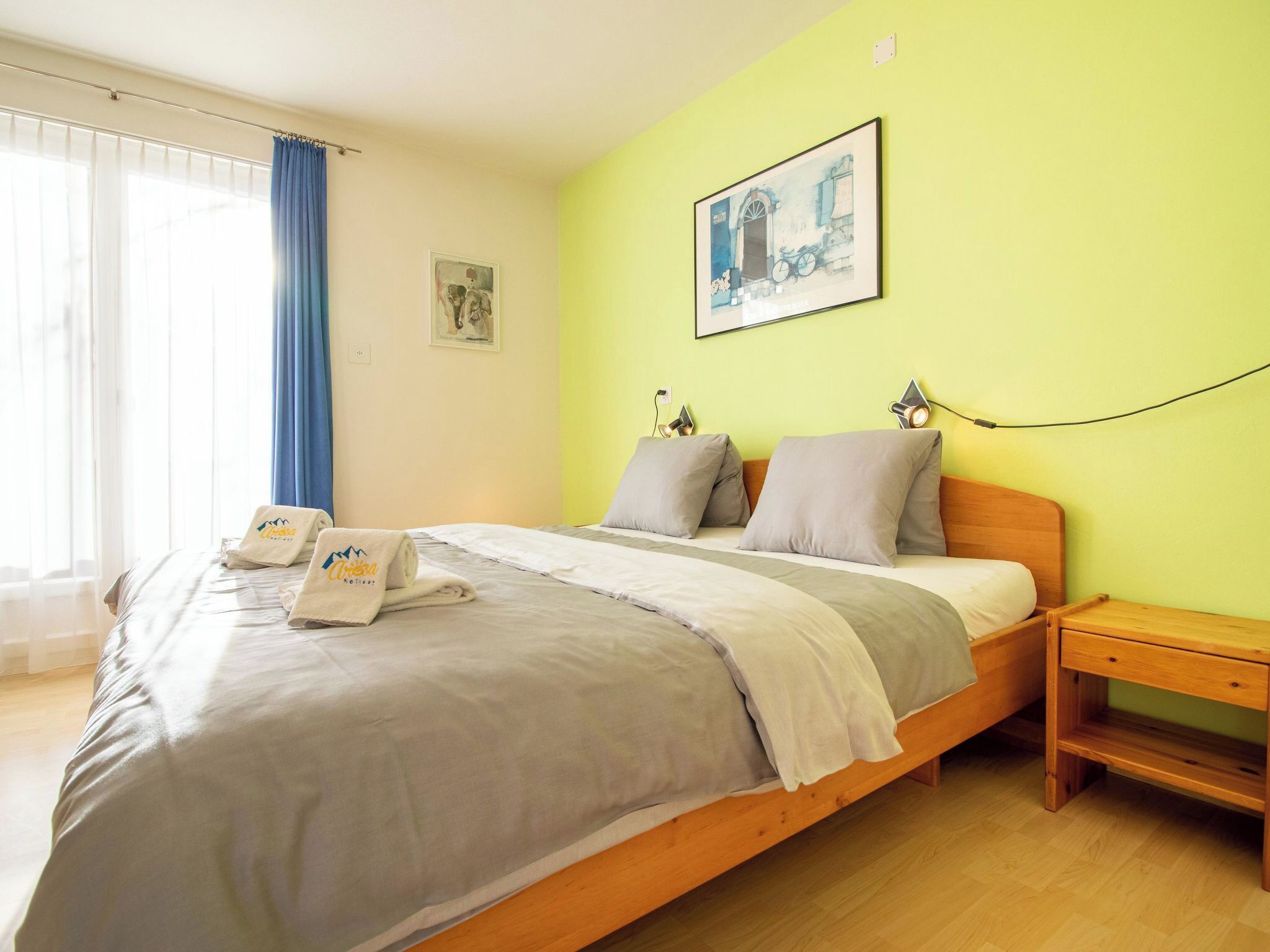 Una cama individual con estructura de madera, una mesita de noche, una lámpara de pared y una ventana con cortinas azules.