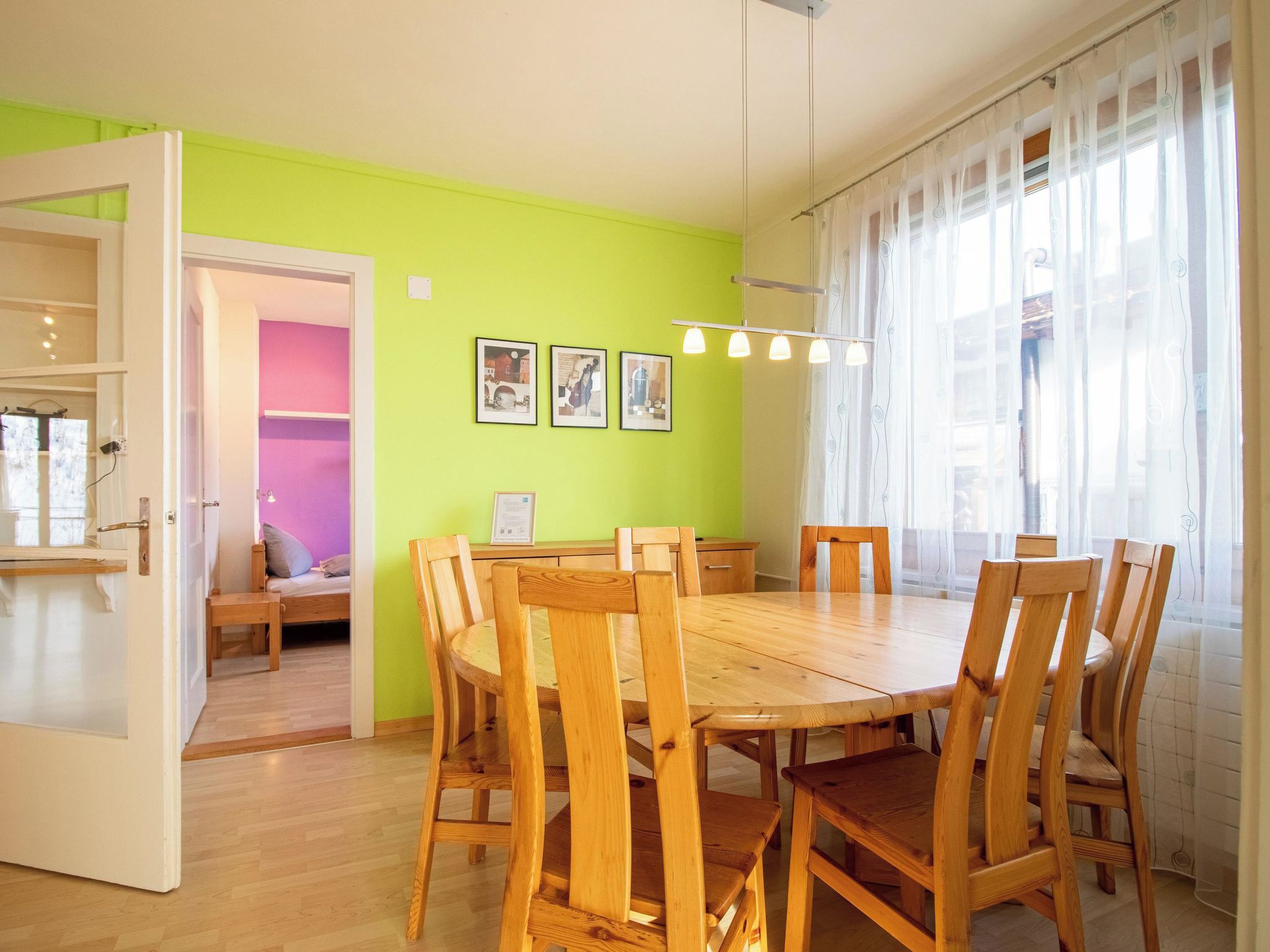 A dining area with a wooden table and chairs, a bright green wall, and a window with sheer curtains.
