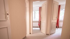 Un pasillo con puertas blancas que dan a habitaciones, suelo alfombrado y una ventana con cortinas rojas.
