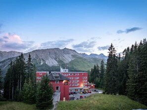 A red building with multiple windows, surrounded by trees and mountains, with a parking area in front. - Chalchboda 1 Appartement/Fewo, Bad, WC, 6-Bettwohnung by Interhome (Arosa)