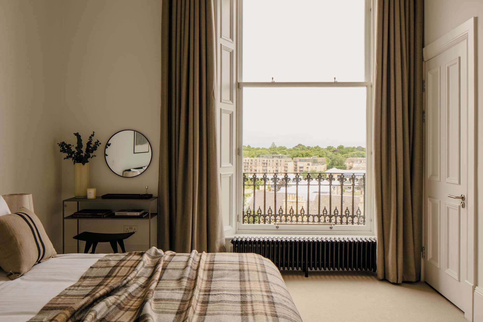A bedroom with a bed, a bedside table, a mirror, and a window with a view of buildings.